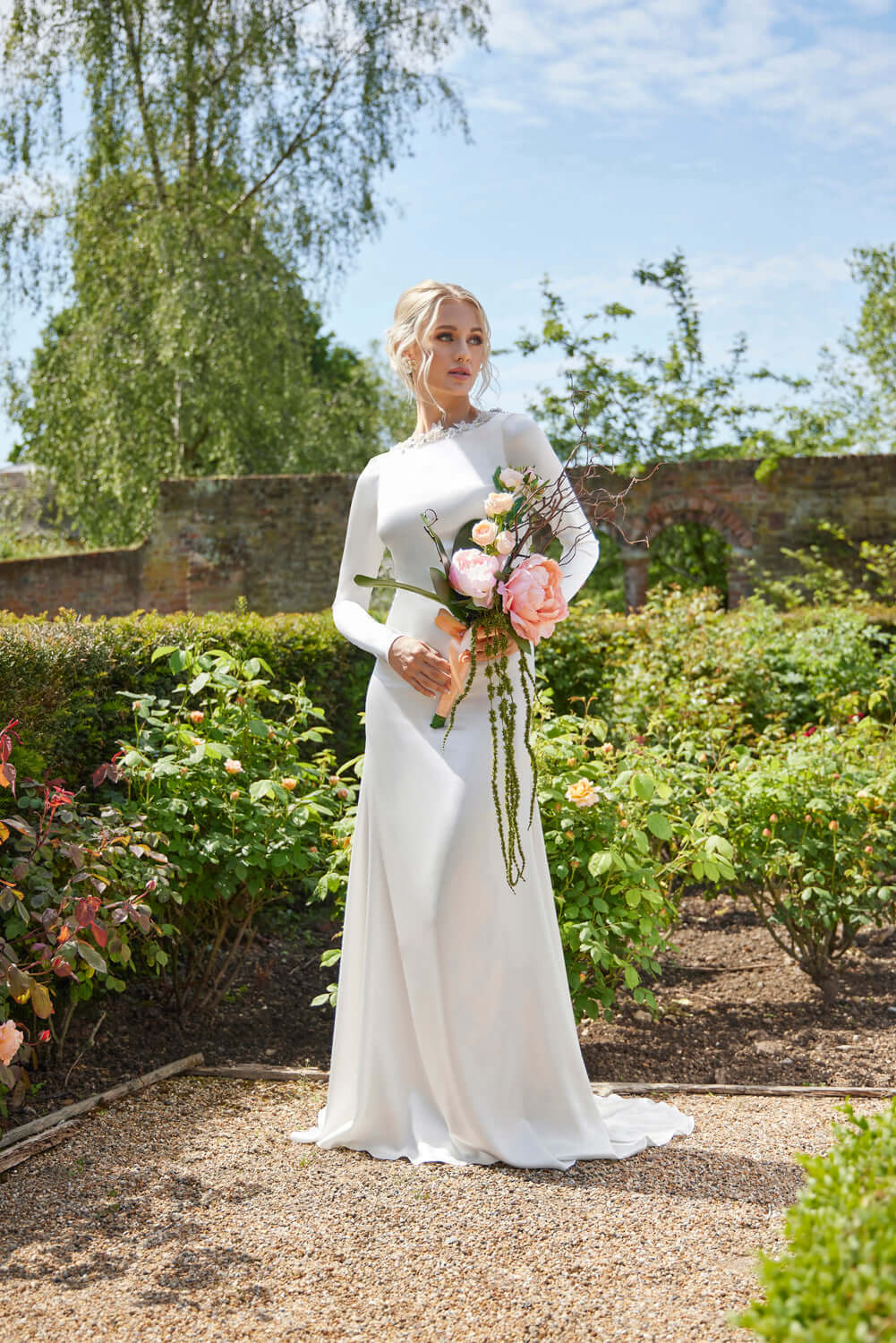 Kassia Wedding Dress - Elegant Silk Gown with Crystal worn by bride in garden holding bouquet of flowers.