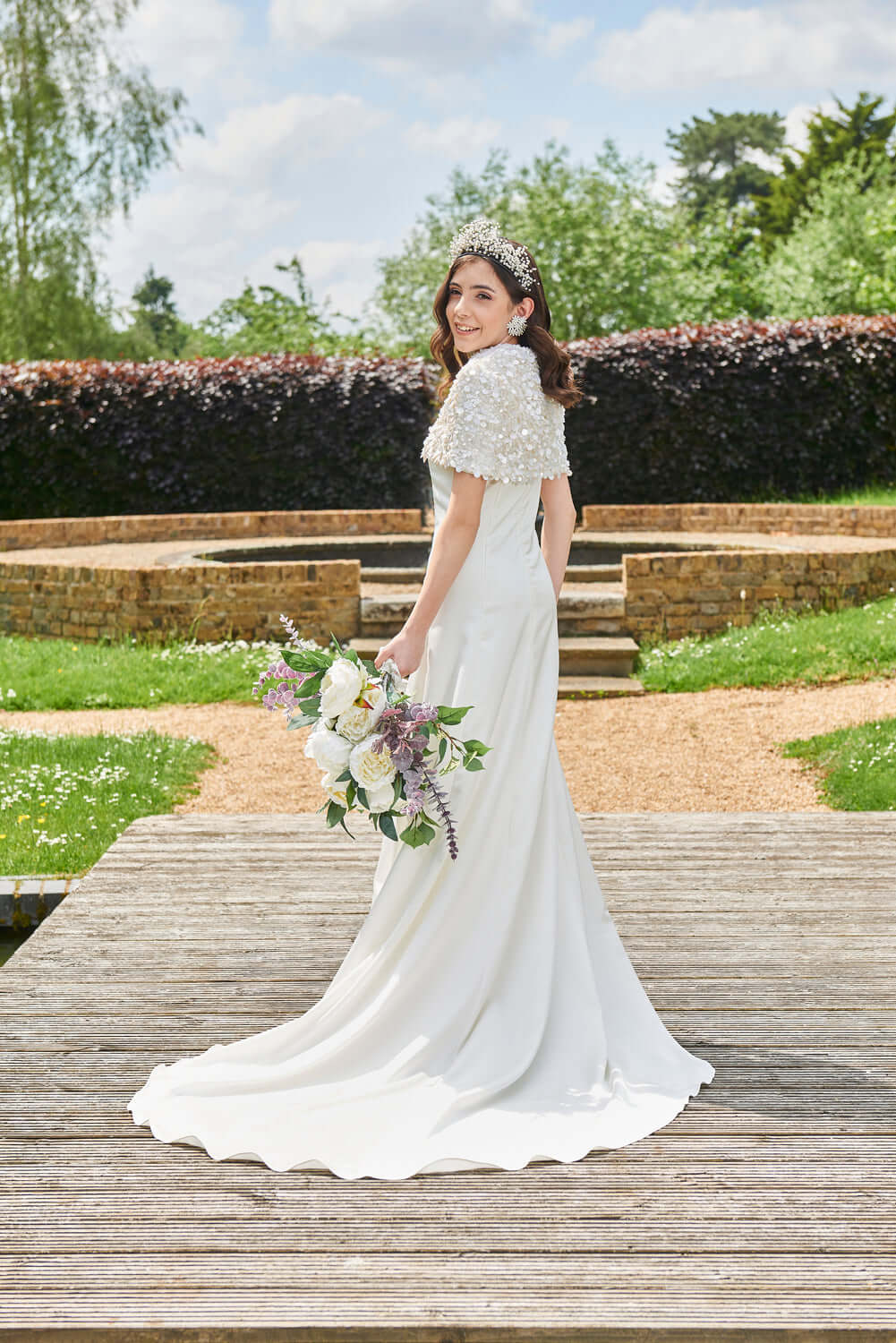 Daneriss Bridal Cape - White Sequined Short Cape worn by bride on a garden path holding a bouquet.