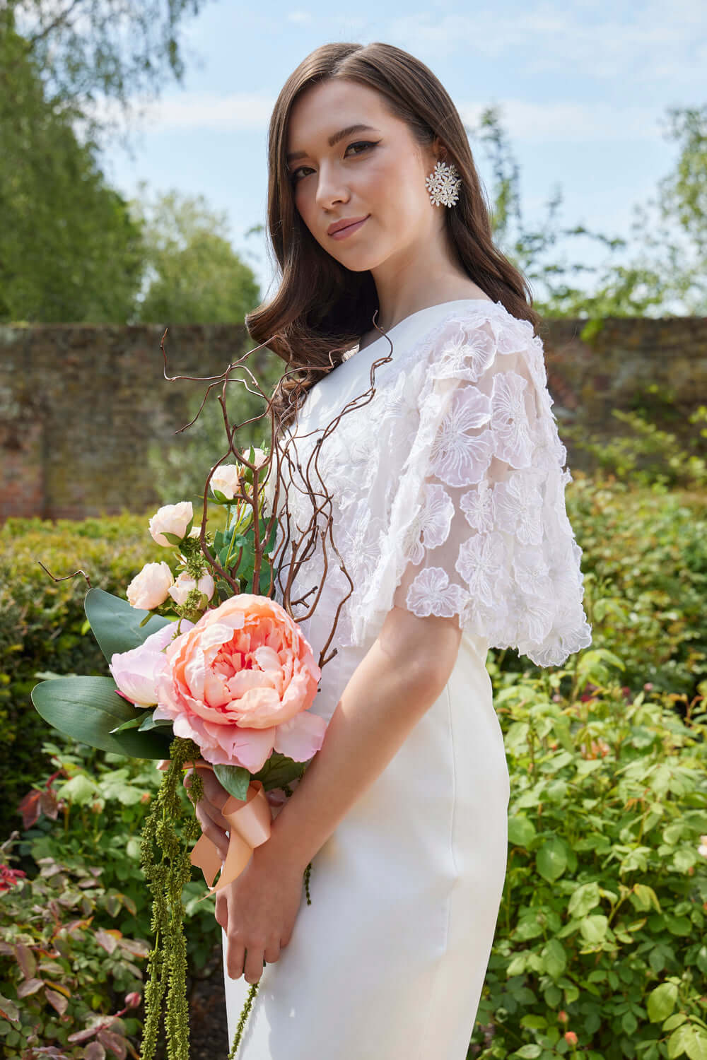 Gilia Midi Wedding Dress with Embroidered Sleeves model holding bouquet in a garden setting by Eve Couture London.